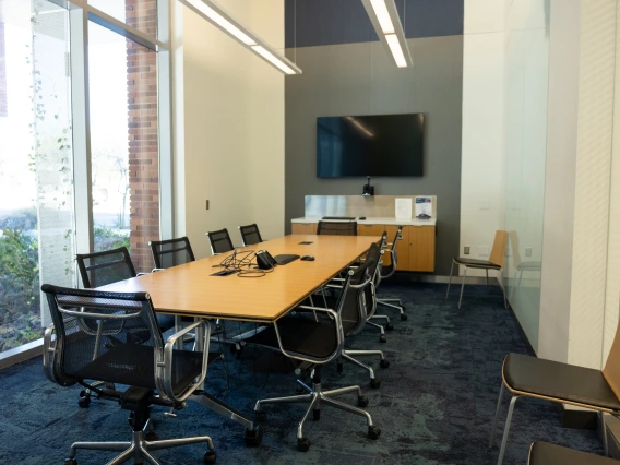 A modern conference room with a large table, several chairs, and a TV screen on the wall. Bright, natural light enters through floor-to-ceiling windows.