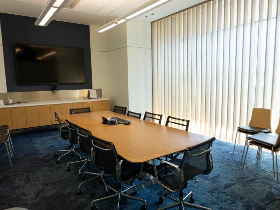 Modern conference room with a large table, several chairs, a TV screen mounted on the wall, and a window with vertical blinds.