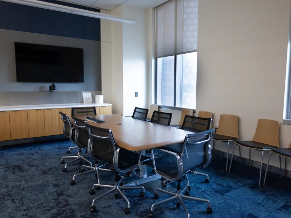 Modern conference room featuring a large wooden table, surrounded by swivel chairs, and equipped with a widescreen TV on the wall.