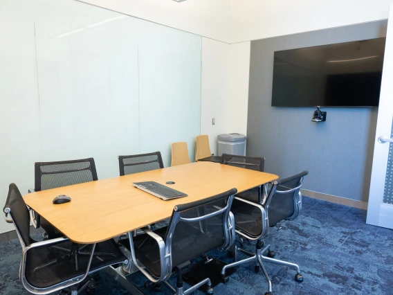 Modern conference room featuring a large wooden table with several chairs, a TV mounted on the wall, and a glass whiteboard. No logos or company names visible.