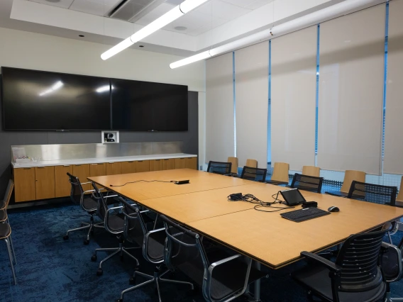 A modern conference room featuring a large table with chairs, a wide-screen monitor on the wall, and a counter with supplies.