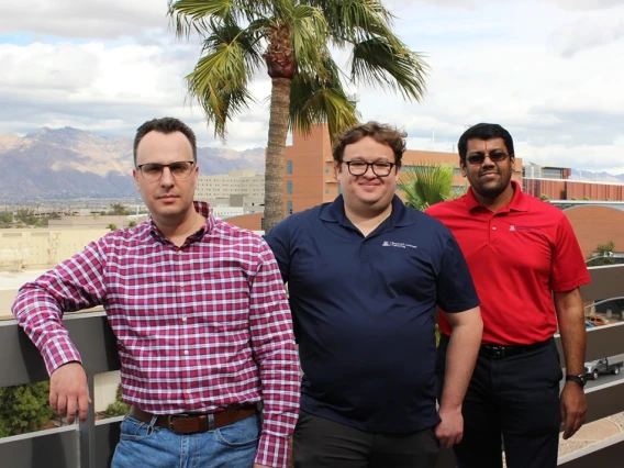 Three individuals standing on a balcony with a backdrop of mountains and a palm tree. They are dressed in casual business attire.