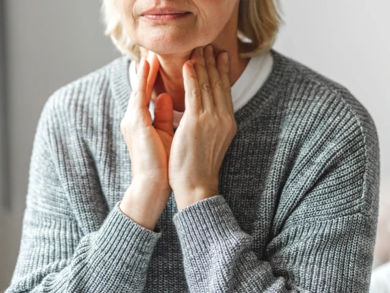 Cropped image of a person holding their throat, wearing a gray sweater over a white shirt.