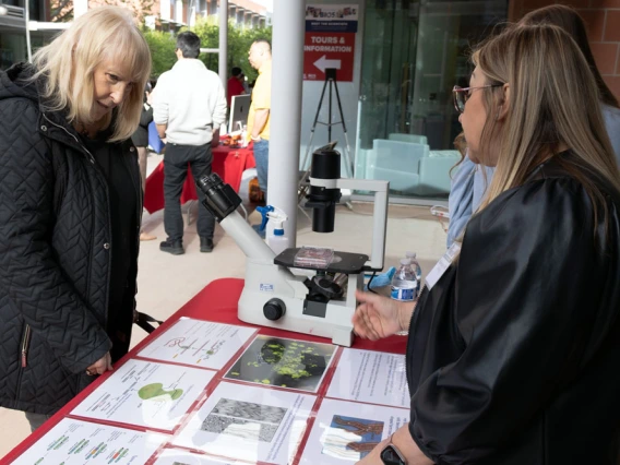 Two individuals are engaged in conversation at an information booth, which features a microscope and various educational materials on display, including maps and images of cells at an outdoor public event.