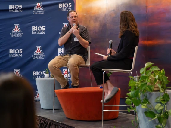Two individuals participating in a discussion at the University of Arizona's BIO5 Institute. One is seated on a stool and speaking into a microphone, while the other listens attentively. The backdrop features the institute's logos and an abstract art piece.