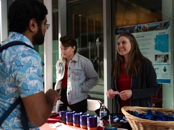 Three individuals engaging in conversation at an informational booth with brochures and promotional items on the table.