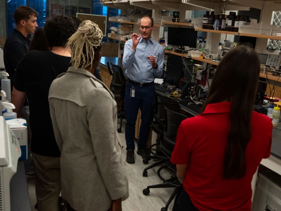 A group of people listening to a scientist in a laboratory filled with equipment.