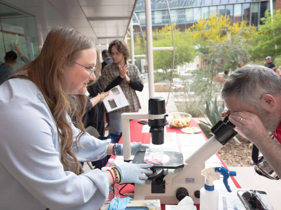Two individuals using a microscope at an outdoor science exhibit, with one person assisting the other in viewing a sample. There are trees and a building in the background.