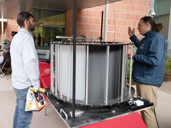 Two individuals conversing beside a large cylindrical scientific apparatus at a public display.