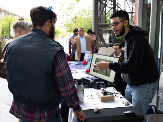A group of individuals engaged in conversation at an outdoor educational booth with various items displayed on the table, including a monitor and some mechanical parts.
