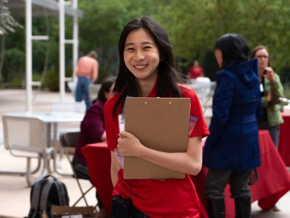 Young person in a red shirt smiling and holding a clip board with an event occuring in the background