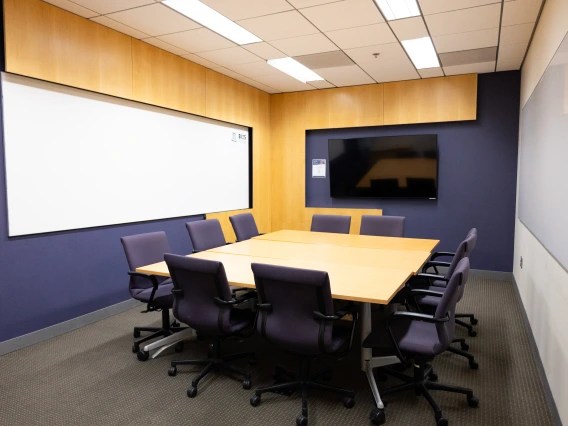 A modern conference room featuring a large wooden table surrounded by black office chairs, a wide whiteboard on one wall, and a flat-screen monitor on the opposite wall.