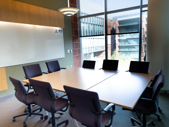 Conference room at BCS featuring a large table with laptops and chairs, adjacent to a whiteboard and large windows overlooking a building exterior.