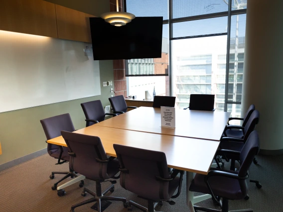 Modern conference room with a large oval table, several chairs, and a wall-mounted monitor, illuminated by natural light from large windows, overlooking an urban landscape.