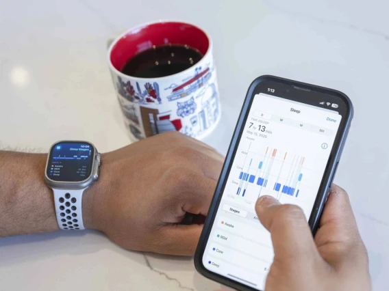 Person checking health statistics on a smartphone with health monitoring app, with an Apple Watch on their wrist and a coffee mug nearby.
