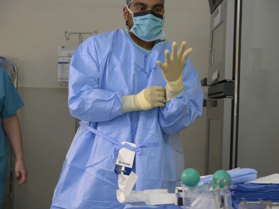 A medical professional wearing scrubs, a surgical mask, and gloves is preparing to put on a second glove in a hospital setting, with another healthcare worker visible in the background.