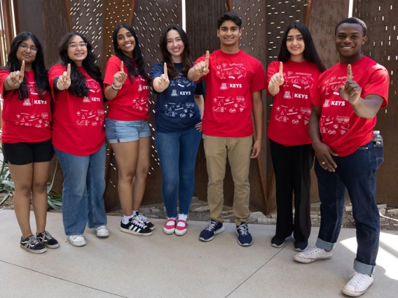 Group of six smiling students in red KEYS t-shirts pointing towards the camera, standing outdoors.