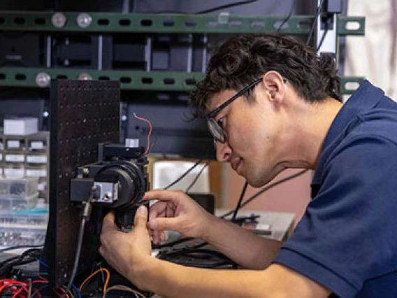 A person is adjusting equipment in a tech workshop, surrounded by shelves filled with various tools and electronic devices.