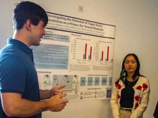 Four individuals are engaged in a discussion in front of a scientific poster at a conference. One person is presenting, pointing towards the poster, as three listeners, including one with a name badge, attentively engage. The content on the poster includes diagrams and text related to neuroscience research.