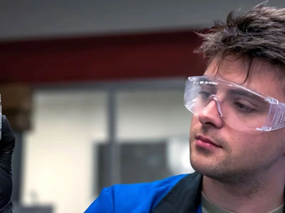 Person wearing safety goggles and gloves holding a chemical bottle in a laboratory setting.