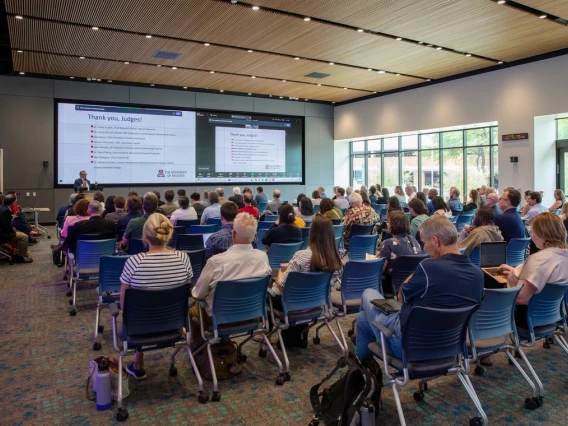 A presentation at a conference with a speaker near a large screen displaying a slide titled "Thank you, Judges!" to an audience seated in rows, taking notes and listening attentively in a modern hall with large windows.
