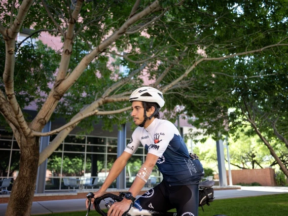 Person in a cycling outfit and helmet sitting on a bike under lush trees, outside a red-brick building.
