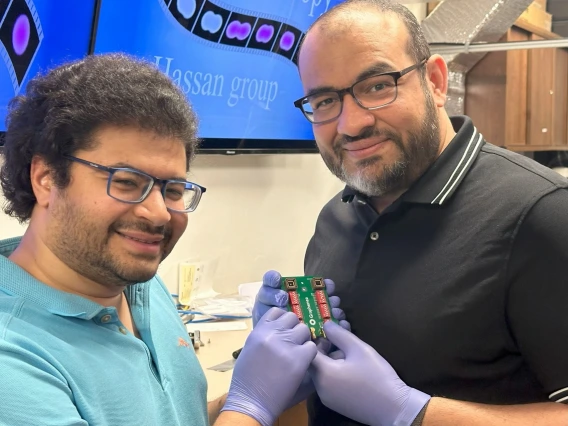 Two individuals in a lab setting, wearing gloves, hold an electronic device, with a screen in the background displaying "Hassan Group".