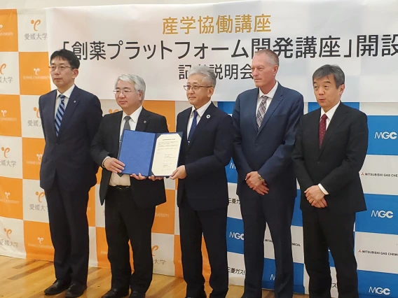 Group of five individuals at a formal event, holding a certificate, with a backdrop featuring logos of Mitsubishi Gas Chemical and other entities.