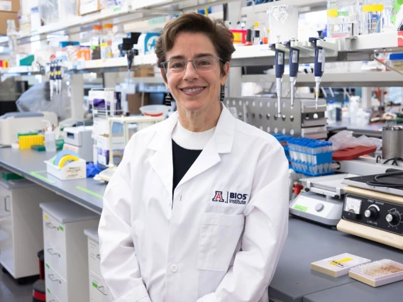 A person wearing a lab coat with a "BIO5" and a University of Arizona logo stands smiling in a laboratory filled with scientific equipment.