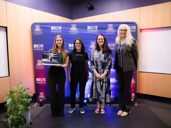 Four individuals smiling and posing together at the University of Arizona's BIO5 Institute, standing in front of a banner with the institute's logo. One person is holding a laptop.