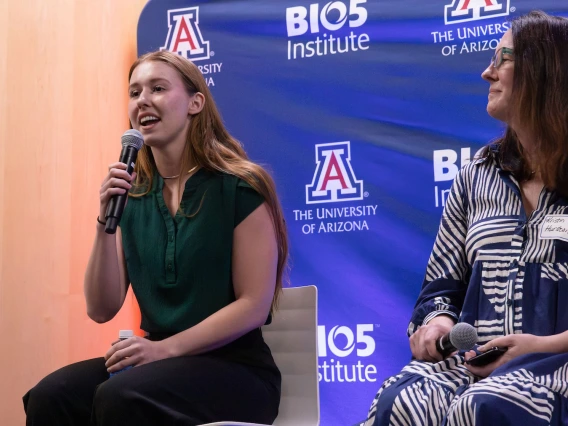 Two individuals seated on stage, speaking at an event at the BIO5 Institute, The University of Arizona. The person on the left holds a microphone and is speaking, while the person on the right listens attentively.
