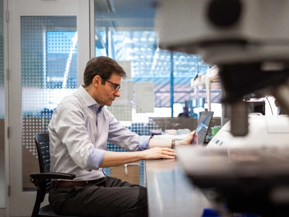 A person wearing glasses is working at a laptop in a modern laboratory, surrounded by scientific equipment.