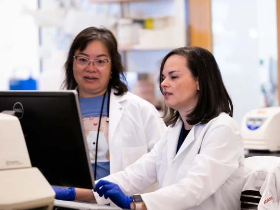 Two scientists in lab coats working together at a computer in a laboratory setting.
