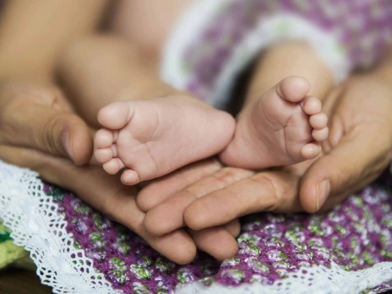 Close-up of an adult's hands gently holding a baby's feet, with both resting on a purple and green knitted blanket.