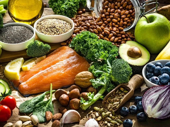 Assorted healthy foods including salmon, nuts, fruits, vegetables, and seeds spread on a wooden surface.