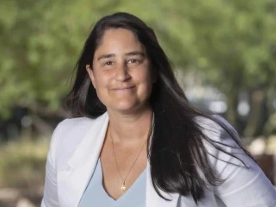 Portrait of an individual smiling at the camera, wearing a white blazer, set against a blurred natural background.
