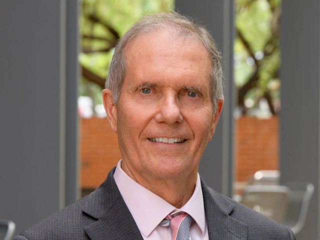 Portrait of person wearing a suit and tie, smiling in front of a blurred background featuring a tree and a building's windows.