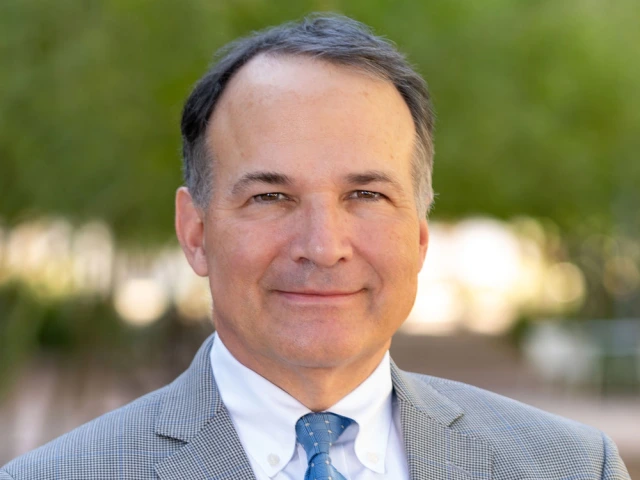 Portrait of a smiling individual in a gray suit and blue tie, outdoors with blurred trees in the background.