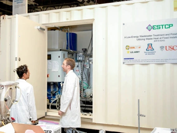 Two individuals in lab coats discussing in front of a large experimental setup labeled as 'ESTCP, A Low-Energy Wastewater Treatment and Potable Reuse System Using Waste Heat at Installations', featuring logos of the U.S. Army, NAVFAC, USAF, USMC, and USC.