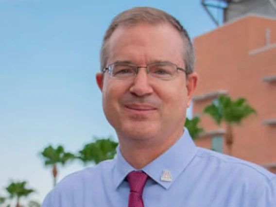 Portrait of a smiling individual wearing glasses, a blue shirt, and a red tie, with a blurred background of palm trees and a building.
