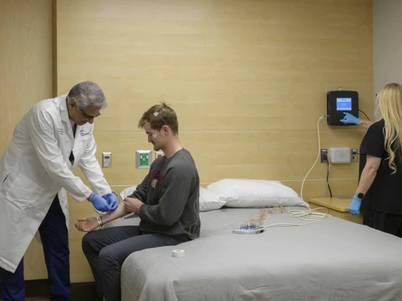 Doctor in a white coat and gloves applying electrodes to the arm of a patient in a medical examination room, with an assistant standing by.