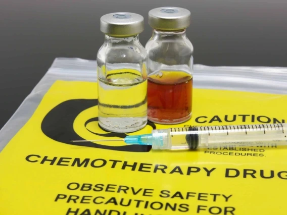 Two vials of chemotherapy drugs, one clear and one amber-colored, beside a syringe on a yellow caution mat labeled "Chemotherapy Drugs: Observe Safety Precautions for Handling."