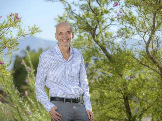 A person standing outdoors, smiling, with blooming trees and a mountainous backdrop. The individual is wearing a patterned blue shirt and gray pants.