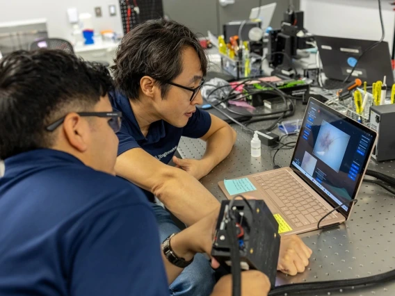 Two individuals examining data on a laptop in a lab with various scientific instruments and equipment around them.