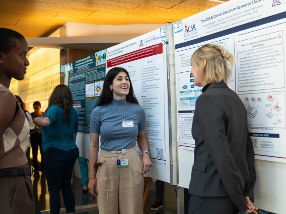 Two individuals discussing research posters at an academic conference, one smiling and engaging with the other who is attentively listening. The backdrop features detailed scientific posters with charts and text.