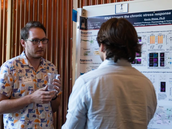 Two individuals engaging in a discussion in front of a scientific poster presentation at a conference. One person is holding a glass of water, and the poster includes various charts and data related to RNA research on stress response.