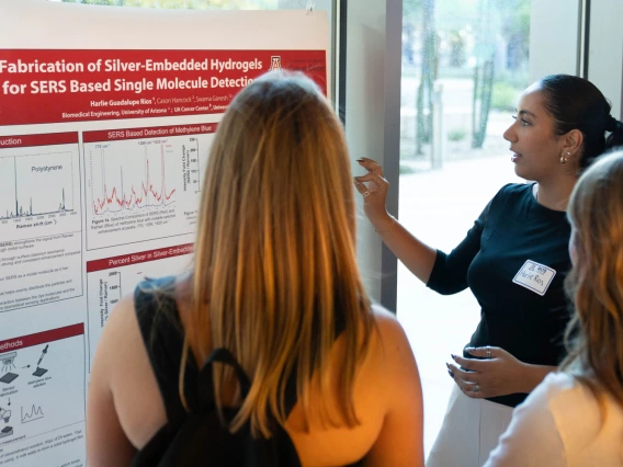 Two individuals examine a research poster titled "Fabrication of Silver-Embedded Hydrogels for SERS Based Single Molecule Detection" at a University of Arizona event. One individual is presenting, pointing at a section of the poster, while the other listens attentively.