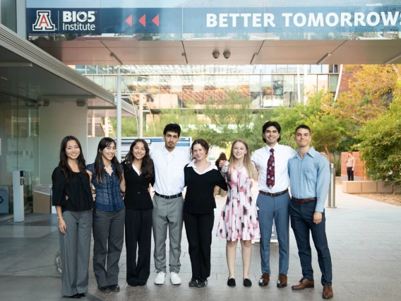 A group of eight diverse individuals smiling and posing in front of the BIO5 Institute, under a sign that says "BETTER TOMORROWS". They are dressed in business casual attire.