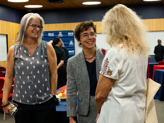 Three individuals engaging in a cheerful conversation at a professional event, with snacks visible on a table in the background. 