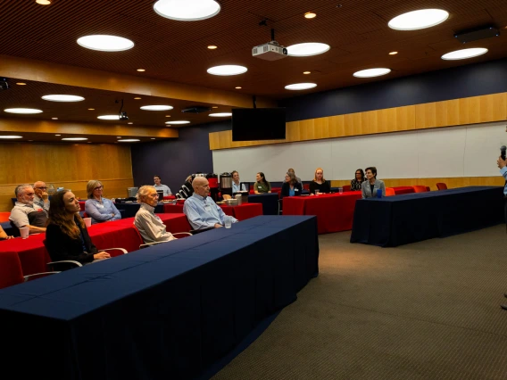 A presenter stands at the front of a lecture hall addressing an audience seated in red chairs. The room is well-lit, with a modern design.